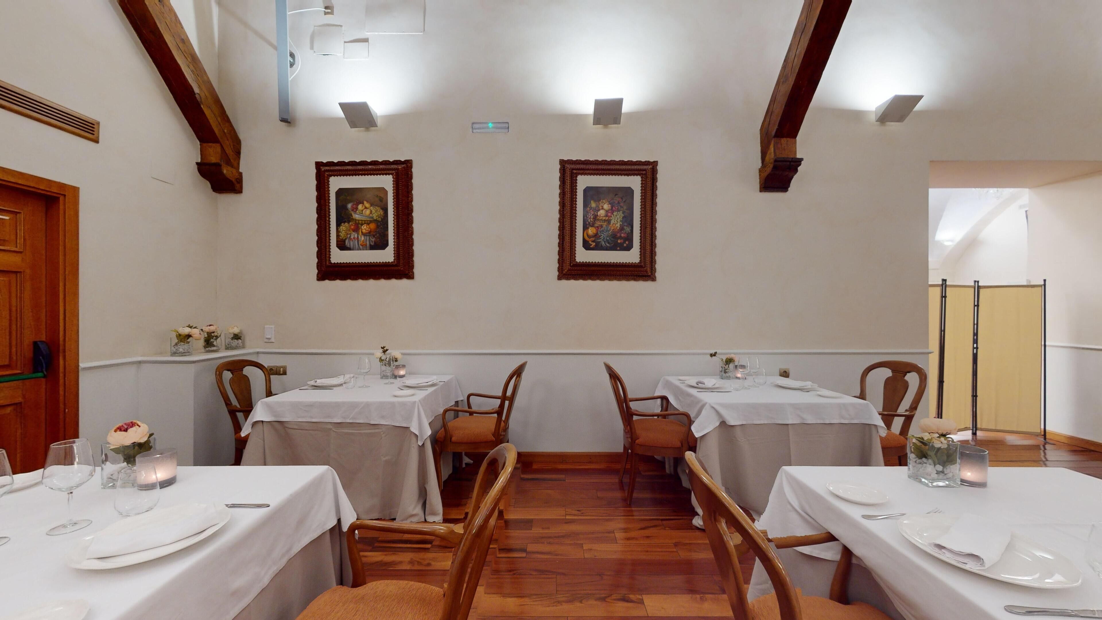Un comedor con cuatro mesas, cada una con mantel blanco, platos y vasos. La sala tiene vigas de madera en el techo y dos cuadros en las paredes.