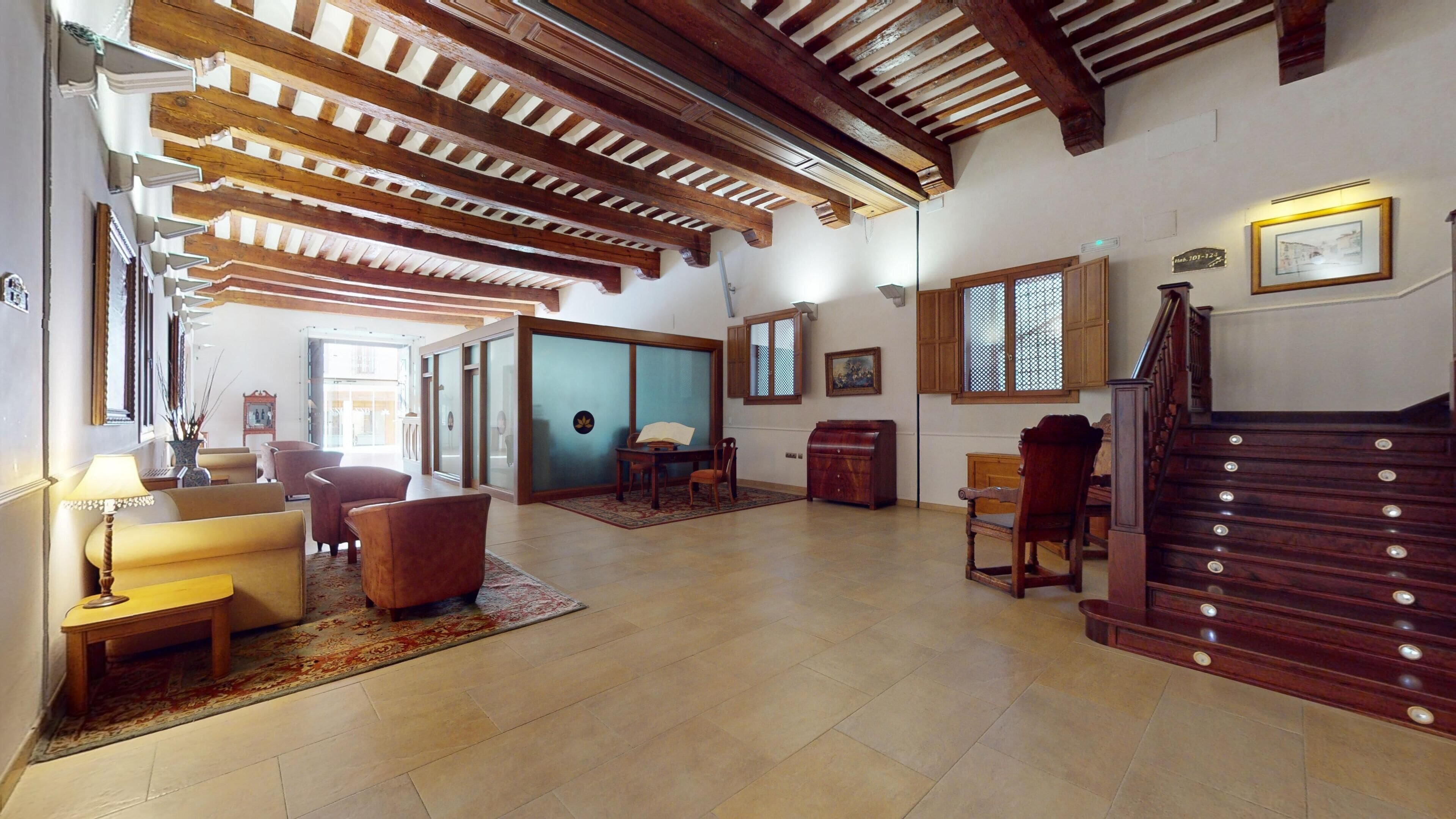 A spacious living area with a high ceiling, wooden beams, a staircase, and a view of the outside.