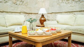 Disposição para o pequeno-almoço numa mesa de madeira com várias frutas e um copo de sumo de laranja, um vaso com plantas verdes e um candeeiro entre dois sofás brancos em frente a uma parede com textura.