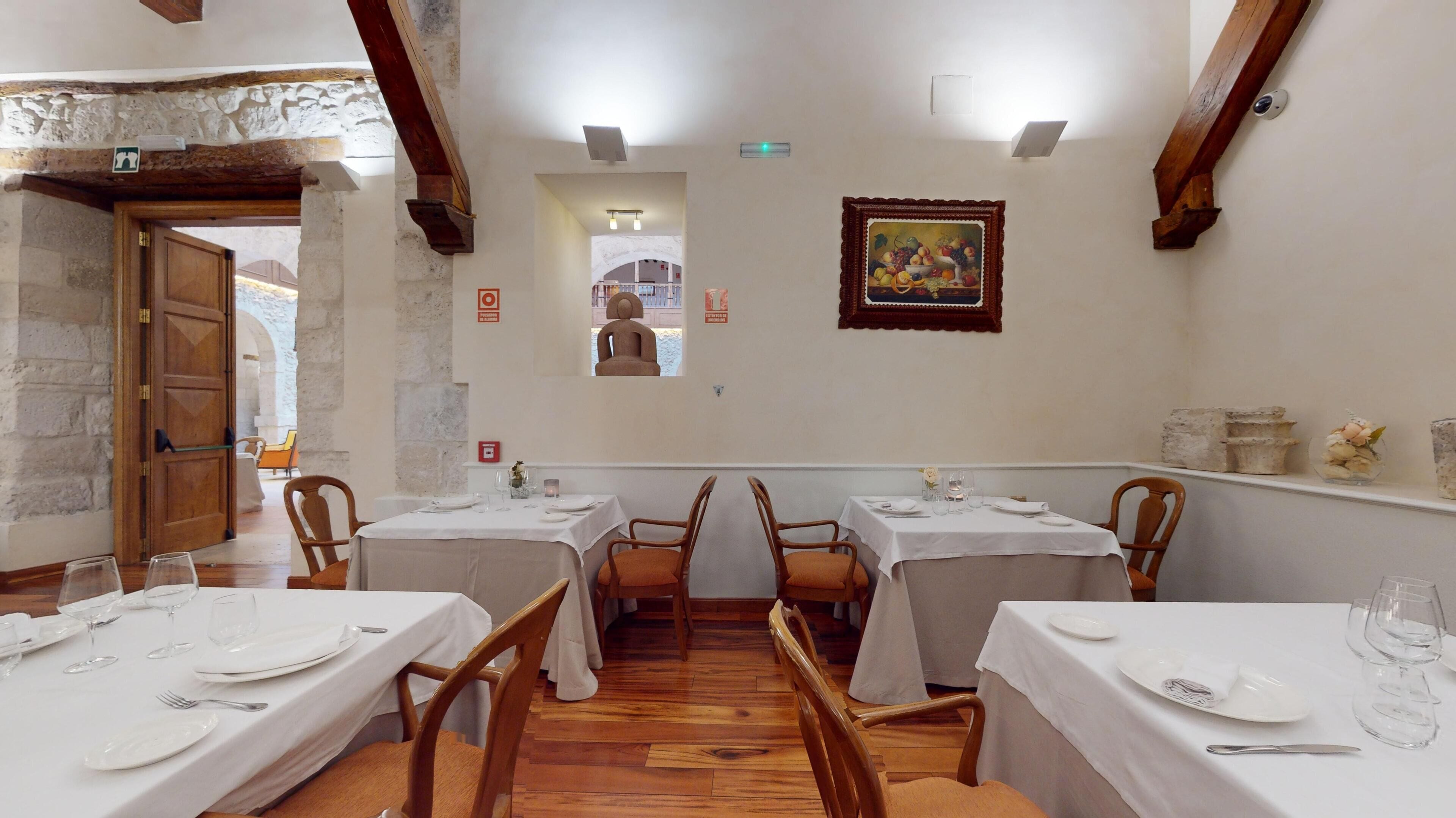 A dining area with tables set for four, wooden chairs, white tablecloths, and a painting on the wall.