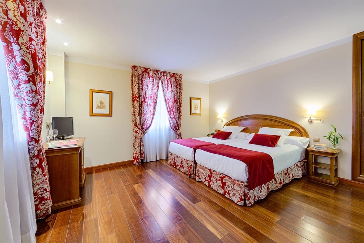 Habitación de hotel con piso de madera, cama con ropa de cama roja y blanca, mesita de noche con lámpara y televisor sobre un mueble de madera.