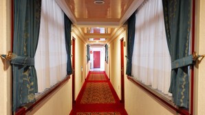 Un pasillo de hotel con alfombra roja, cortinas azules con motivos florales y una puerta roja al final.
