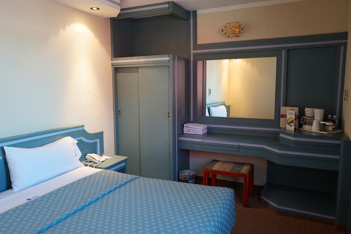 Ein Hotelzimmer mit einem Bett, einem Schreibtisch mit Telefon, einem Spiegel und einem kleinen Hocker.