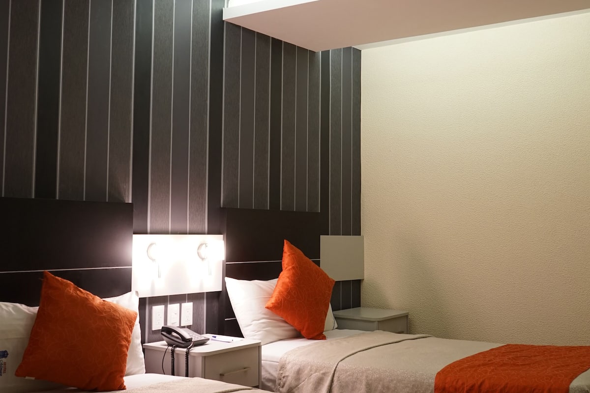 Ein Hotelzimmer mit zwei Betten, jeweils mit einer orangenen Tagesdecke, einem Nachttisch mit Lampe sowie einem großen Fenster mit vertikalen Jalousien.