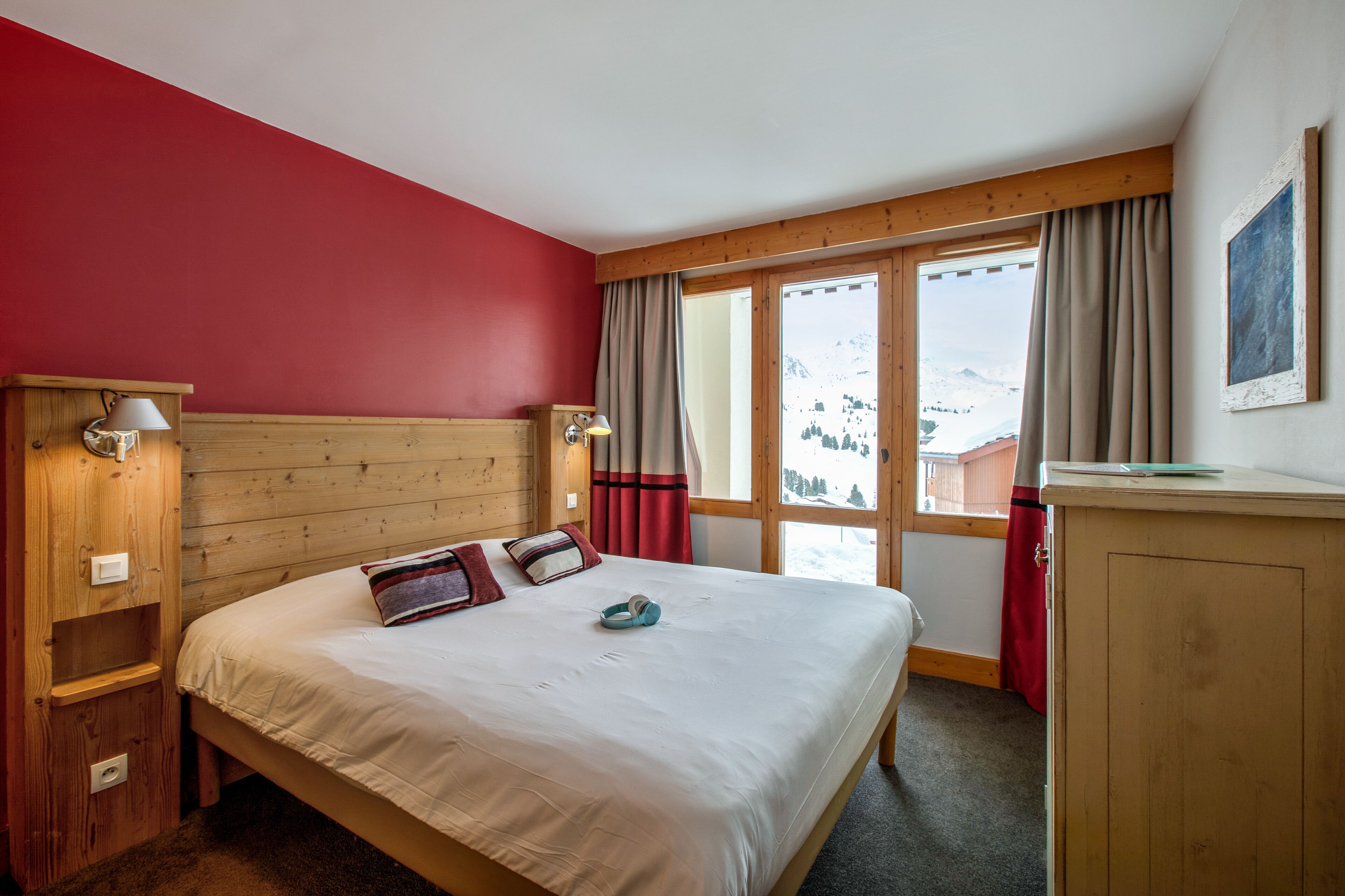 A bedroom with a wooden headboard, a bed with white bedding, a nightstand with a lamp, and a view of a snowy mountain landscape through large windows.