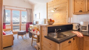 Una cabaña acogedora con interior de madera, una cocina con lavaplatos y cafetera, un comedor con mesa y sillas, y vistas a montañas nevadas a través de amplios ventanales.