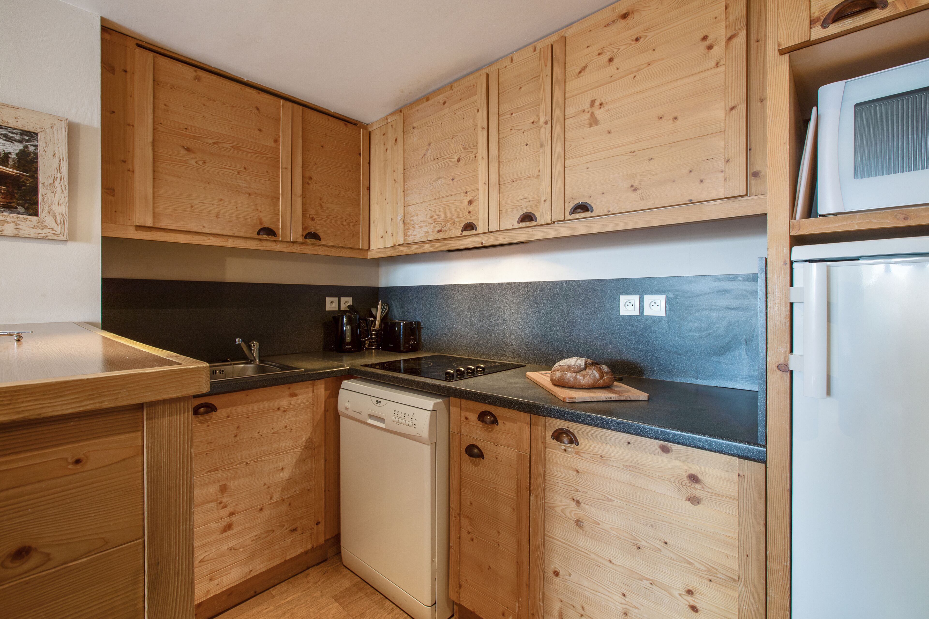 A compact kitchen with wooden cabinets, a dishwasher, a microwave, and a countertop with a loaf of bread.