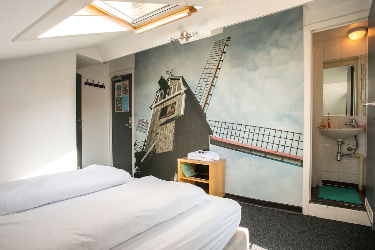 Een hotelkamer met een bed, een klein houten nachtkastje, een muurschildering van een gebouw en een badkamer met een wastafel en spiegel.