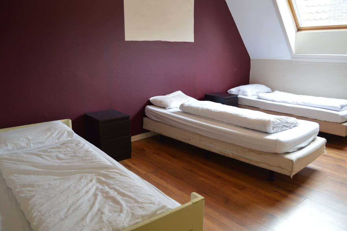 Een kamer met twee enkele bedden, een klein, donker nachtkastje en een raam met uitzicht op de lucht.