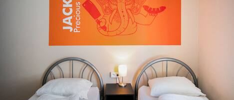 A hotel room with two single beds, a nightstand with a lamp, and a wall-mounted sign reading "JACK'S Precious IPA."