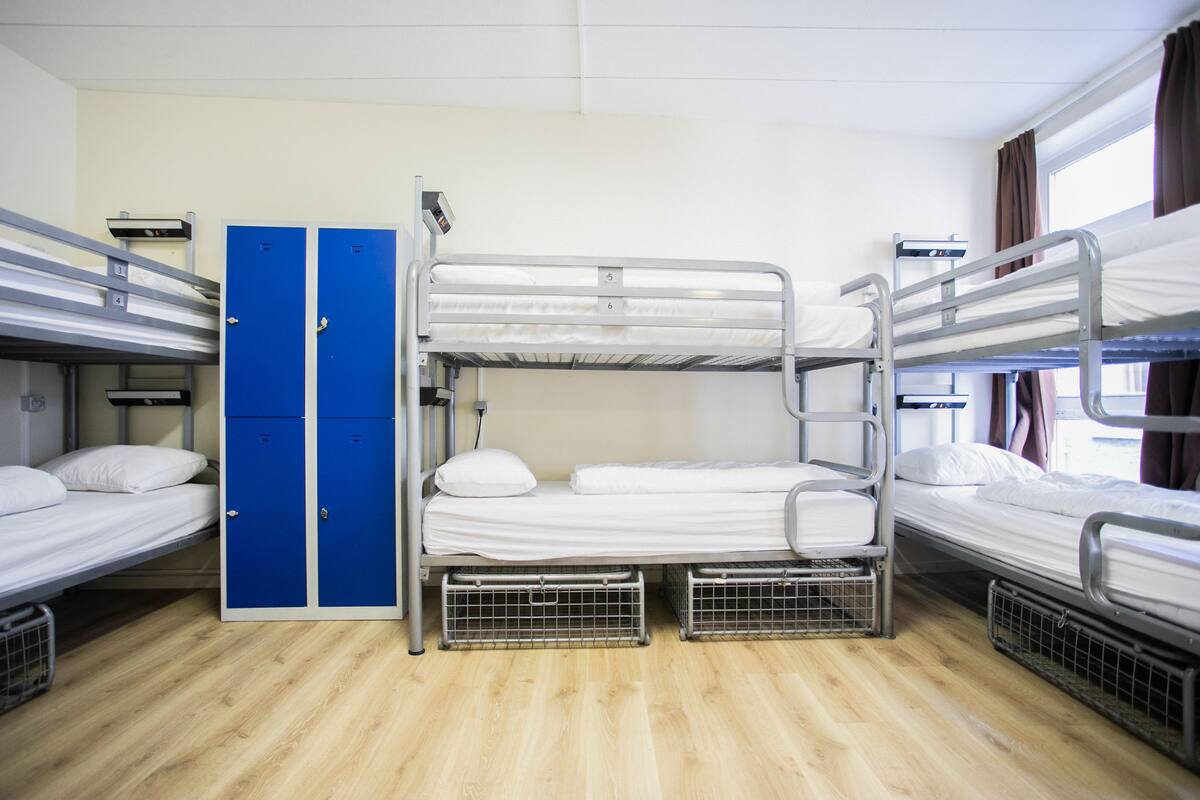 Een studentenkamer met stapelbedden, een blauwe garderobe en een raam met gordijnen.
