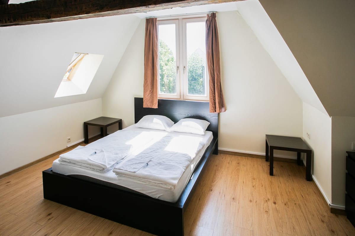 Een slaapkamer met een bed, een raam met gordijnen, een kleine tafel en een nachtkastje.