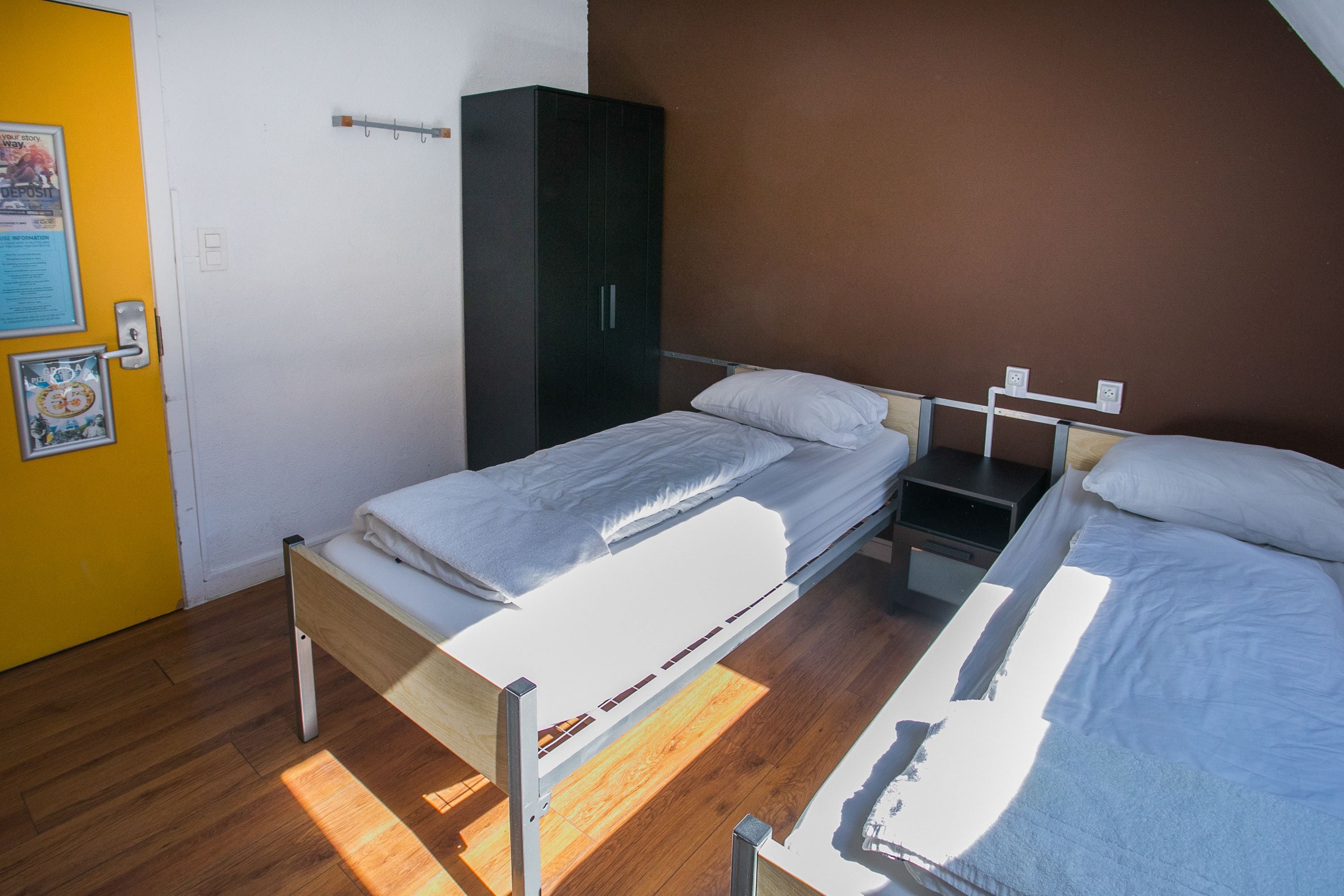 A room with two single beds, a wardrobe, a small table, and a wall-mounted shelf.