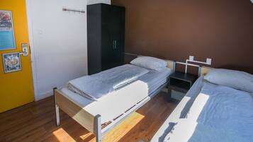 A room with two single beds, a wardrobe, a small table, and a wall-mounted shelf.