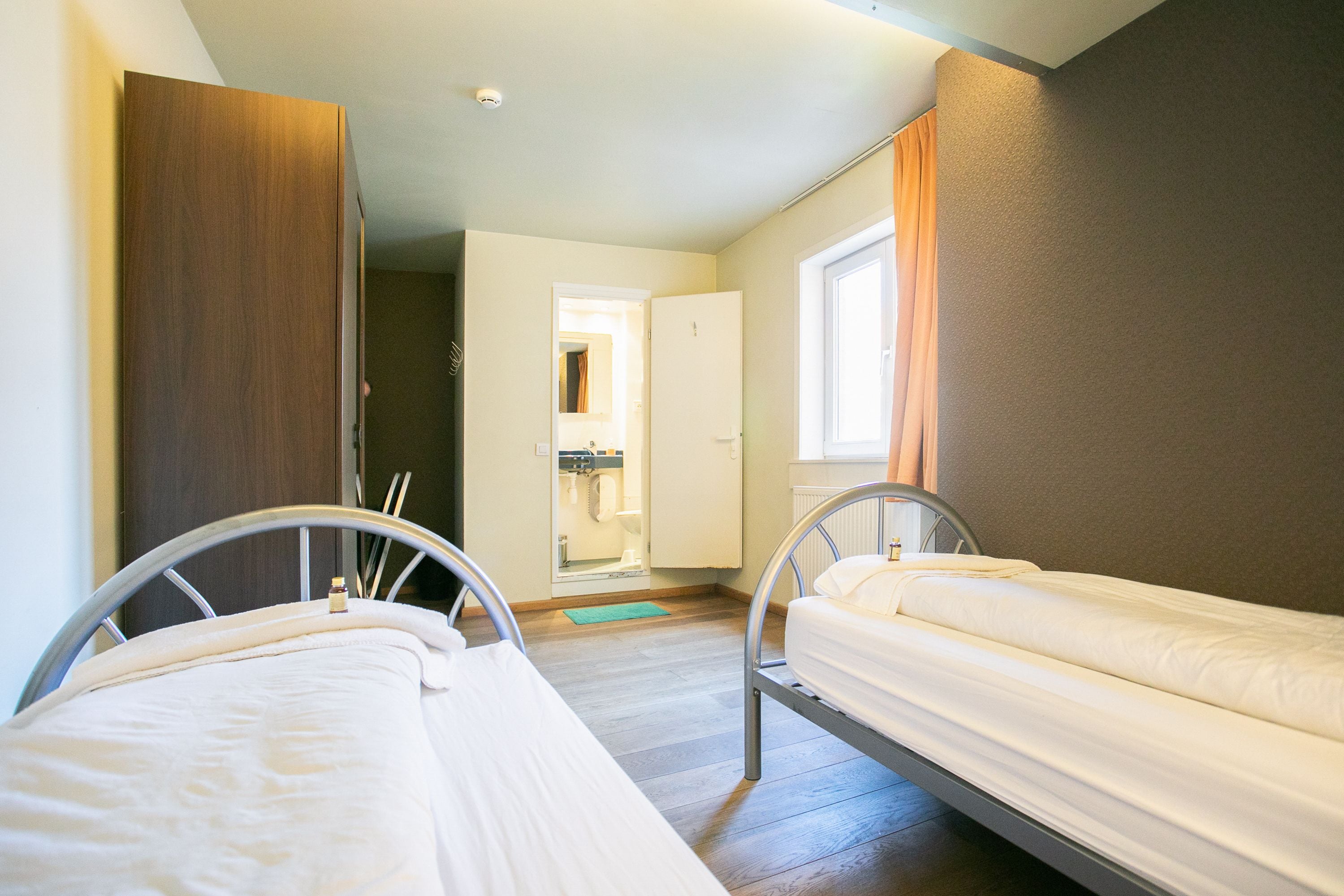 Una habitación pequeña y bien iluminada con dos camas de metal, un armario de madera y un baño visible a través de una puerta abierta.