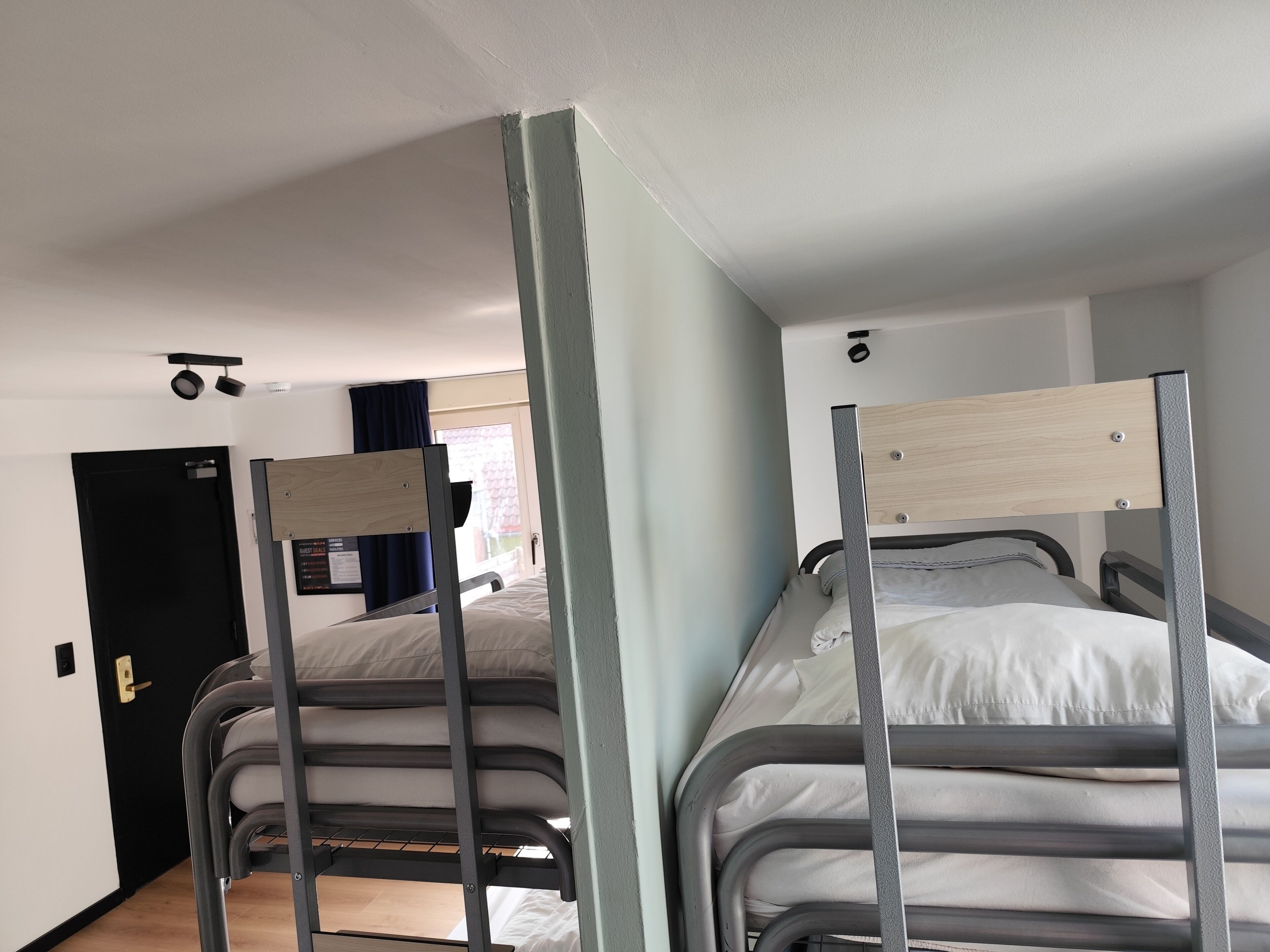A room with two bunk beds, a black door, a window with curtains, and a light switch on the wall.