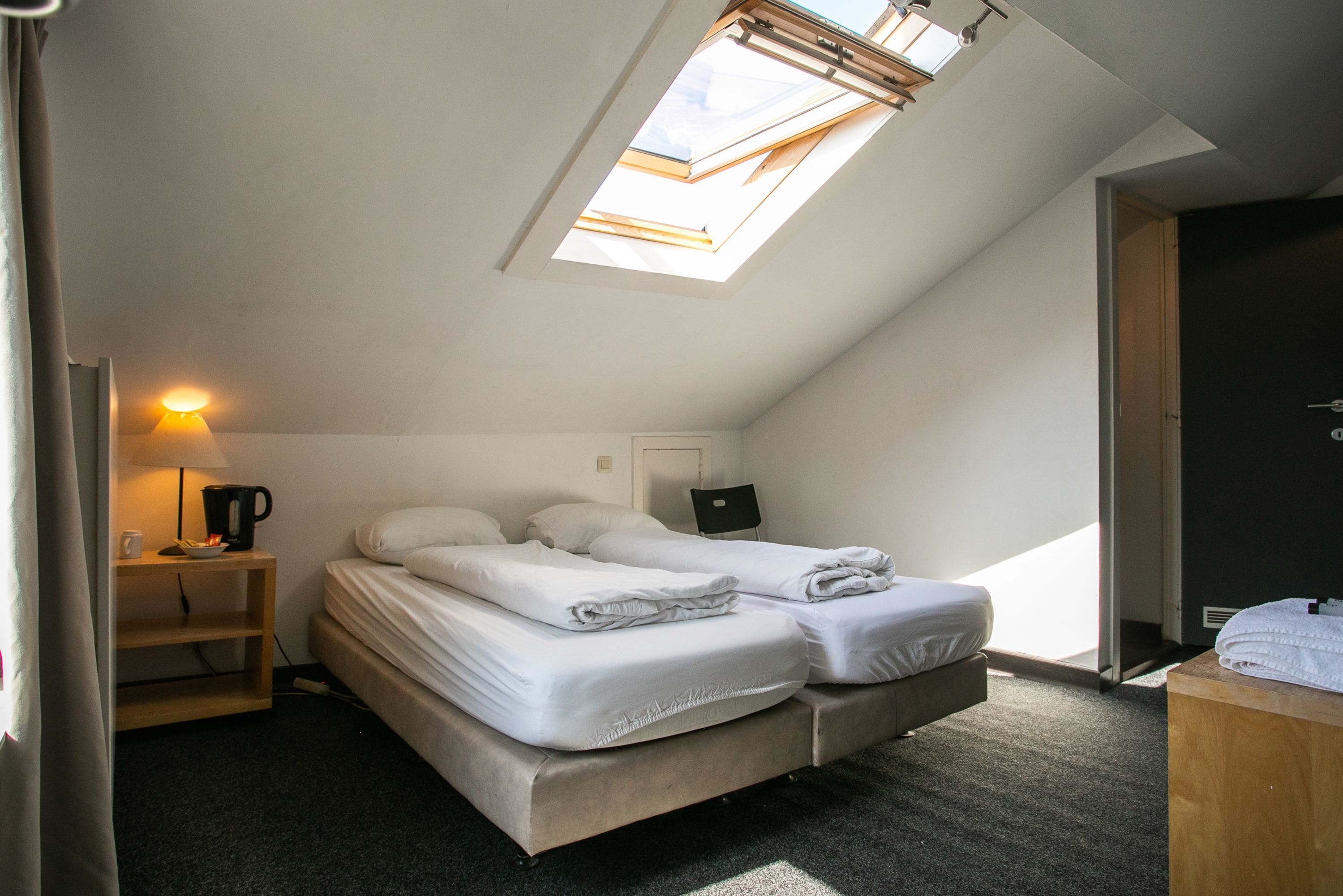 Un dormitorio pequeño y moderno con tragaluz, un escritorio con lámpara y una cama con ropa de cama bien tendida.