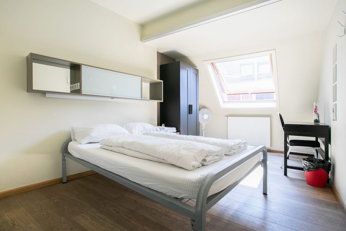 Een enkel bed met metalen frame, een nachtkastje, een stoel, een bureau, een raam en een radiator.
