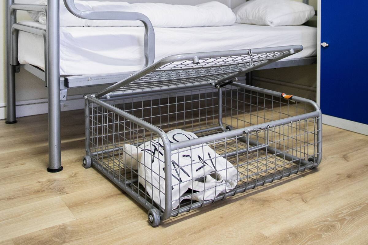 Een stapelbed met metalen frame en witte beddengoed, een blauwe opbergkast en een metalen karretje met een witte tas.