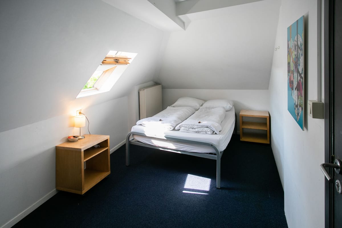 Een kleine, goed verlichte kamer met een eenpersoonsbed, een nachtkastje, een bureau en een raam.