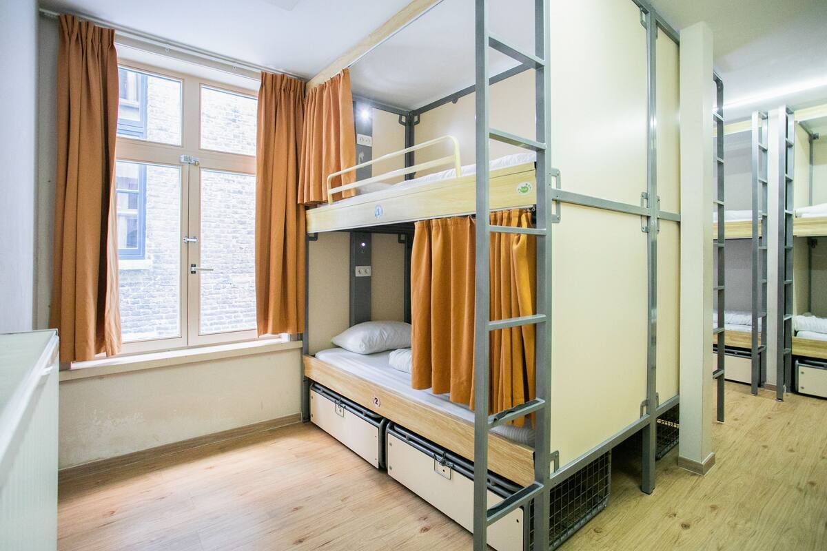Een slaapzaal met stapelbedden, een raam met gordijnen en een bakstenen muur.