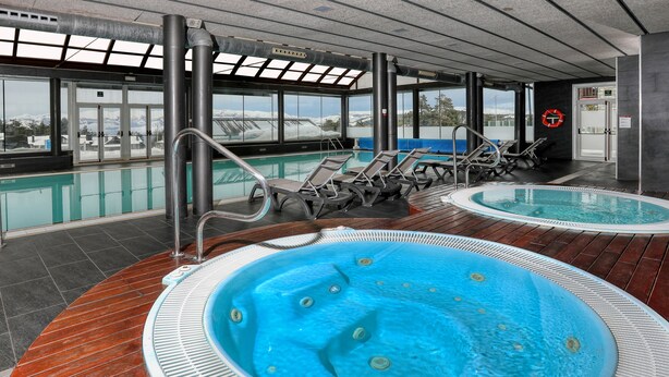 Zona de piscina con dos jacuzzis, sillas de descanso y vistas a montañas nevadas.