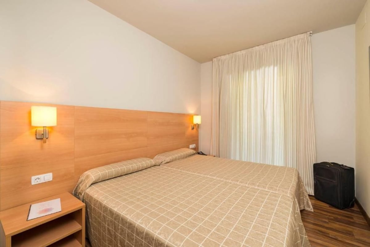 A hotel room with a bed, two bedside lamps, a wooden headboard, a nightstand, a suitcase, and a window with curtains.