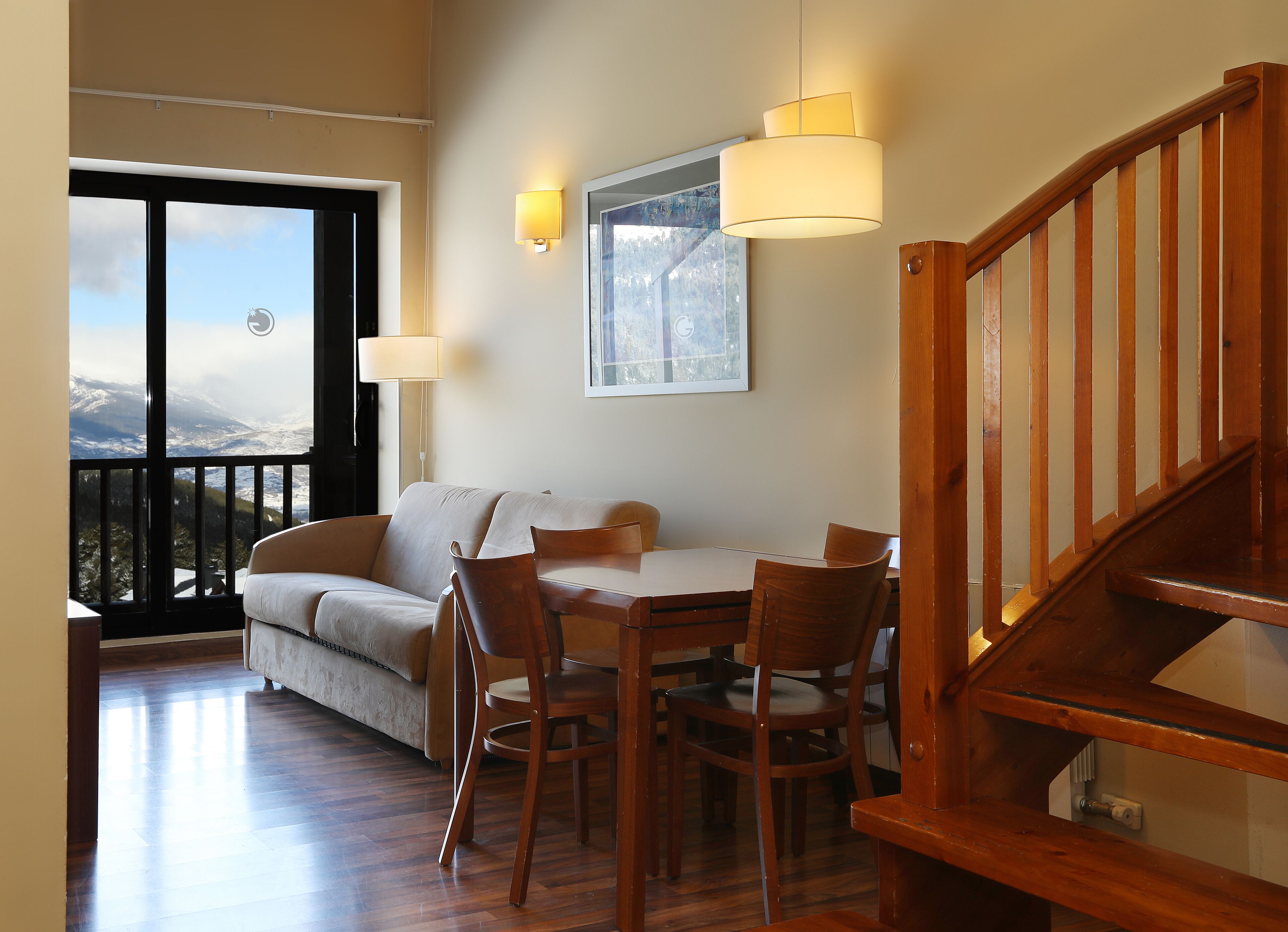 Una sala de estar acogedora con una escalera de madera, un sofá, una mesa de comedor con sillas y un amplio ventanal con vistas a la montaña.