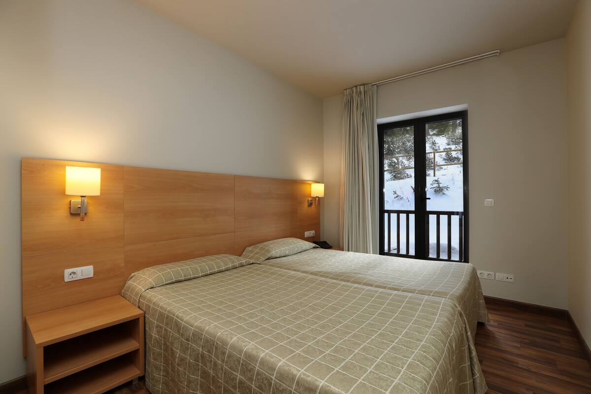 Une chambre d’hôtel avec une tête de lit en bois, deux tables de chevet, un lit recouvert d’une couverture à carreaux et une vue sur un espace extérieur enneigé, visible à travers une porte coulissante.