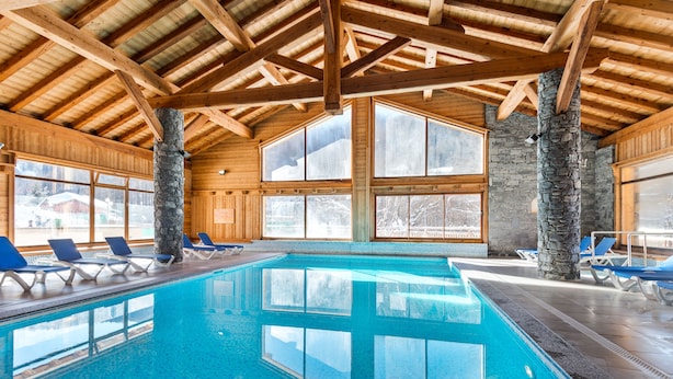 Una piscina interior con agua azul cristalina, vigas de madera, pilares de piedra y sillones azules para relajarse.