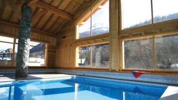 Un espace piscine agrémenté d’un pilier en pierre, de poutres en bois et de grandes fenêtres offrant une vue sur les montagnes.