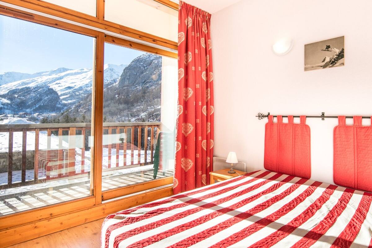 Ein Schlafzimmer mit großem Fenster, einem Bett mit rot-weiß gestreifter Tagesdecke, einem roten Vorhang und einem Bild an der Wand.