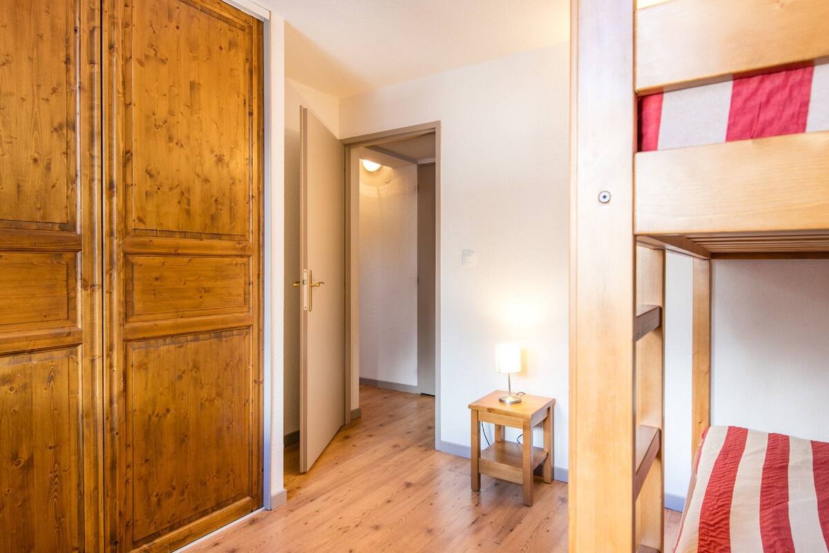 Eine Holztür, ein Korridor mit Holzboden, ein kleiner Tisch mit Lampe und ein Etagenbett mit gestreifter Decke.