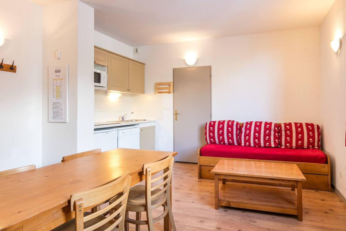 Ein kleiner, gut beleuchteter Raum mit einem Holztisch, Stühlen, einer Couch mit roter Decke und einer kleinen Küchenzeile im Hintergrund.