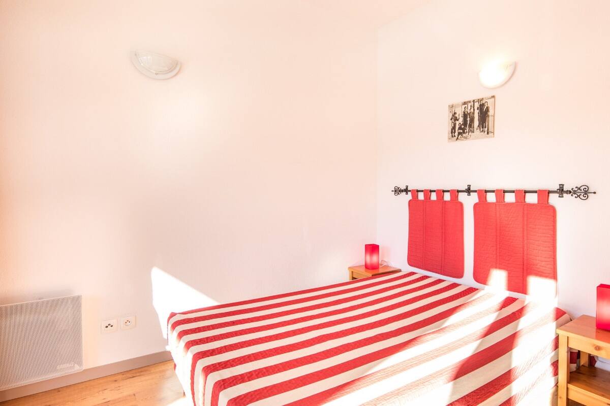 Ein Schlafzimmer mit Bett, Nachttisch, rot-gestreiftem Bettbezug, rotem Vorhang, einem an der Wand hängenden Bild und einer Wandleuchte.