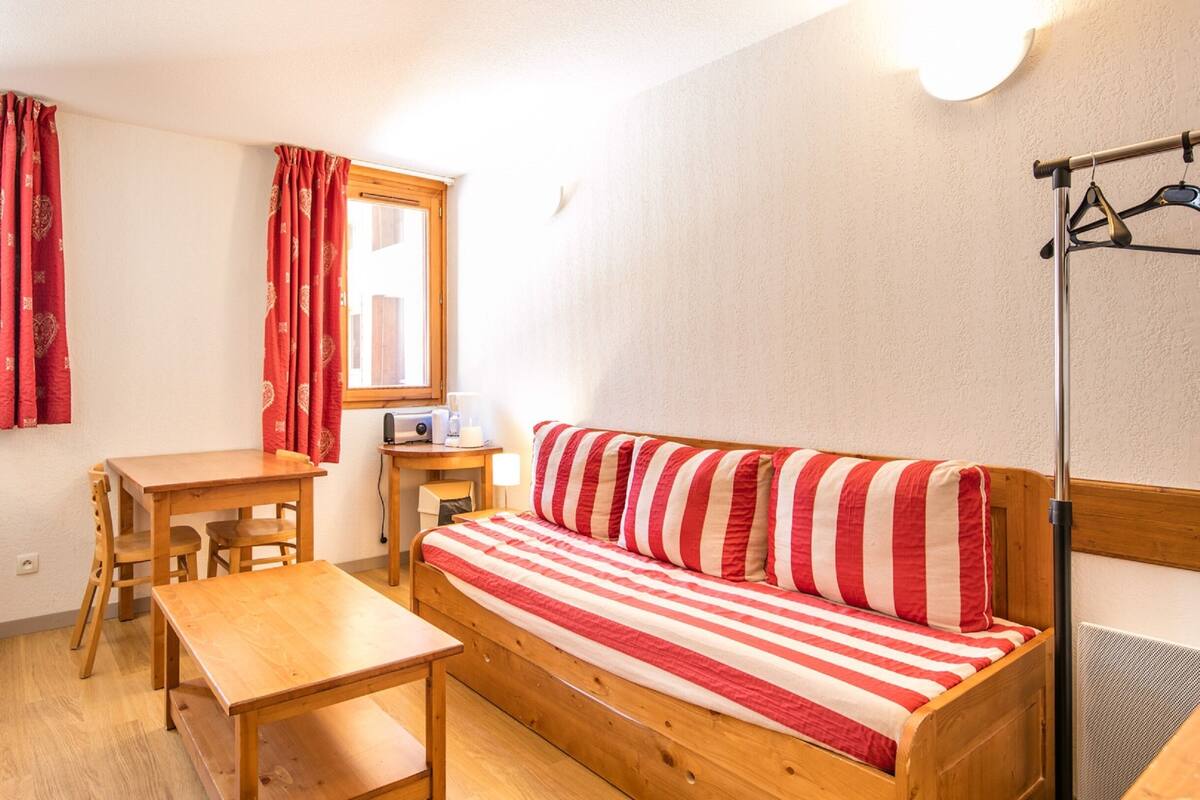 Ein gemütliches Zimmer mit einem hölzernen Bett, einem Tisch aus Holz und einem Stuhl. Es gibt ein Fenster mit roten Vorhängen, eine Heizung und eine Wandleuchte.