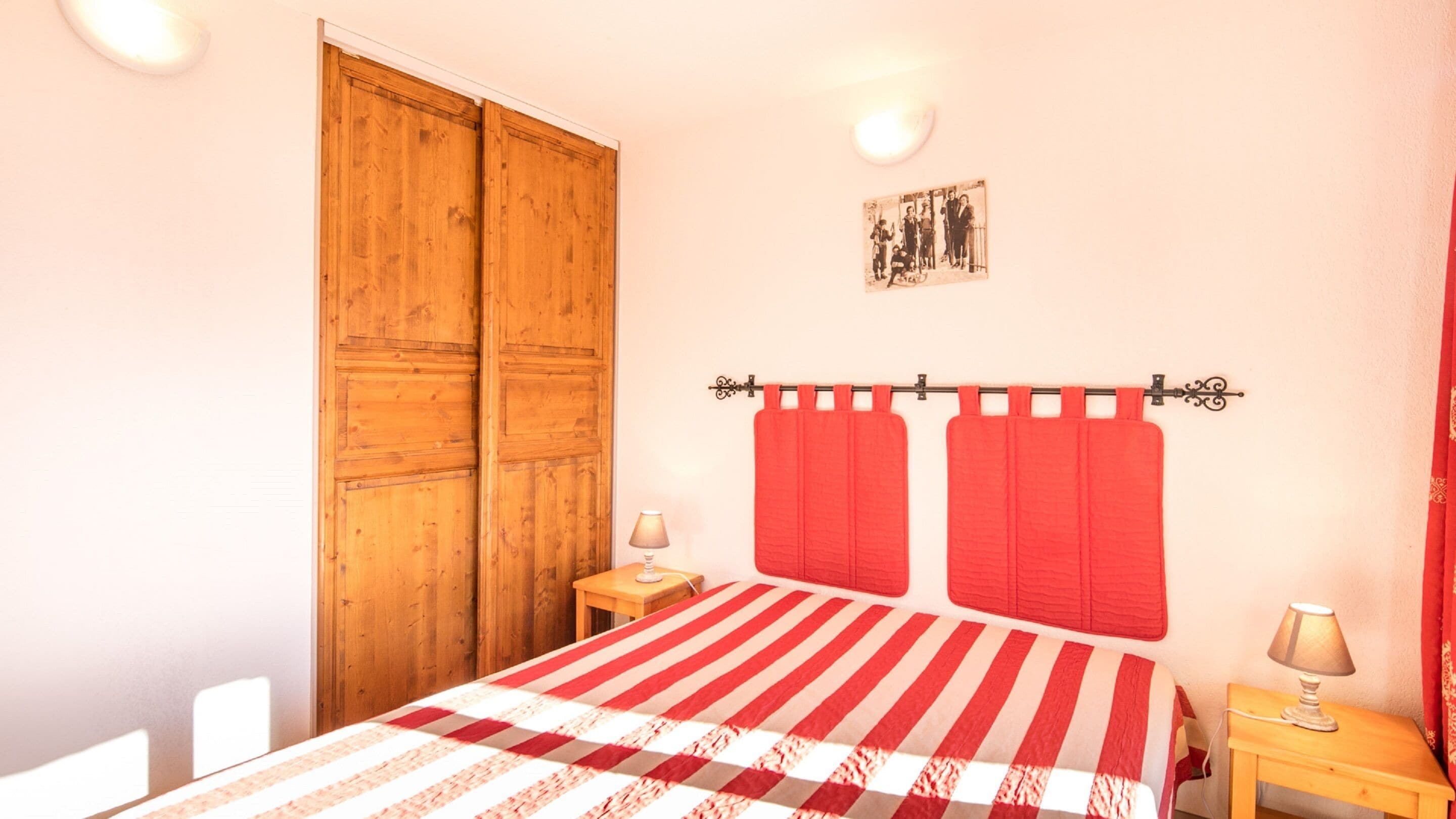 Un dormitorio con cama, armario de madera, dos mesitas de noche, una cortina roja y un cuadro colgado en la pared.