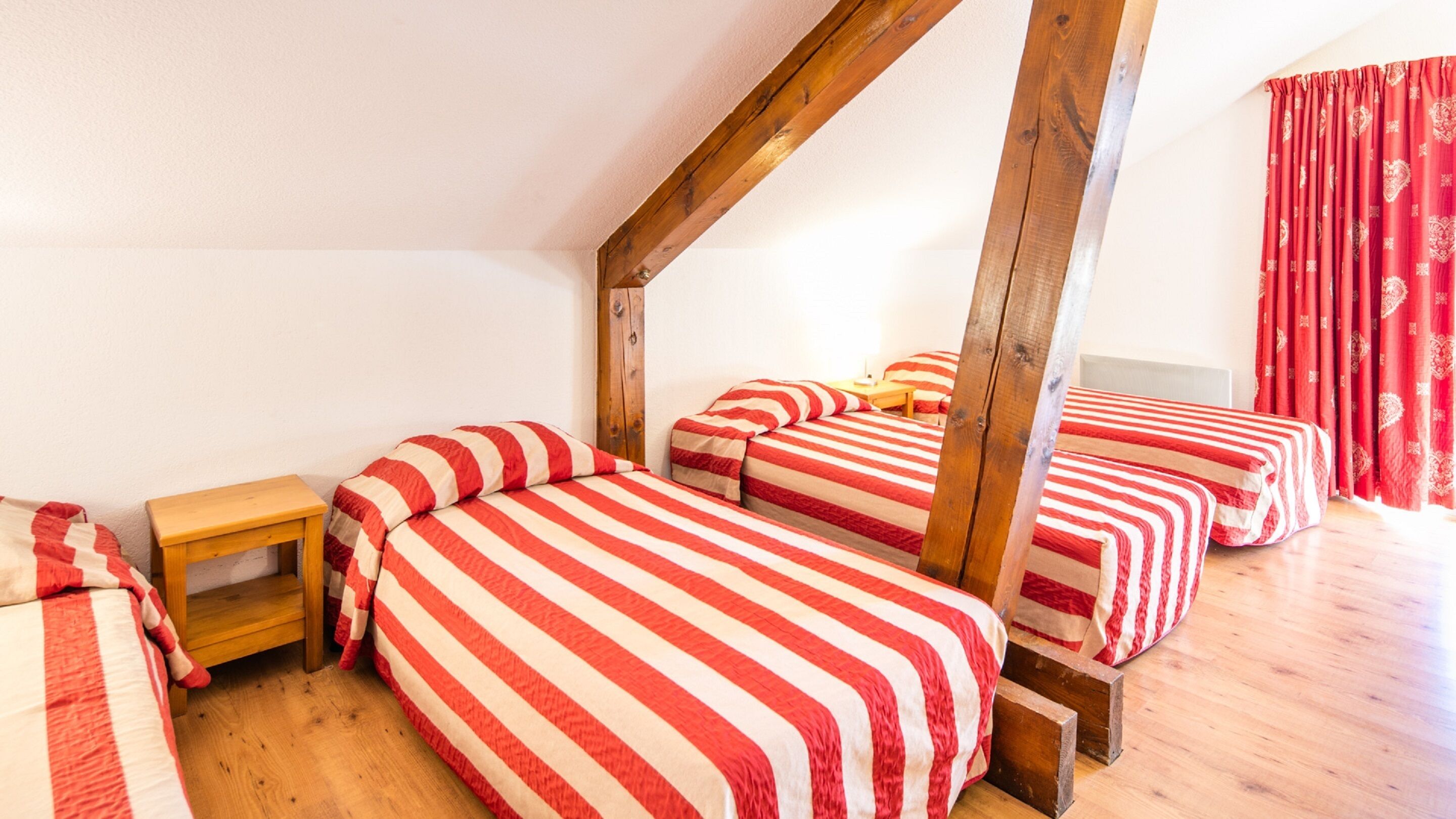 A room with three beds, wooden beams, a small wooden table, and a red curtain.