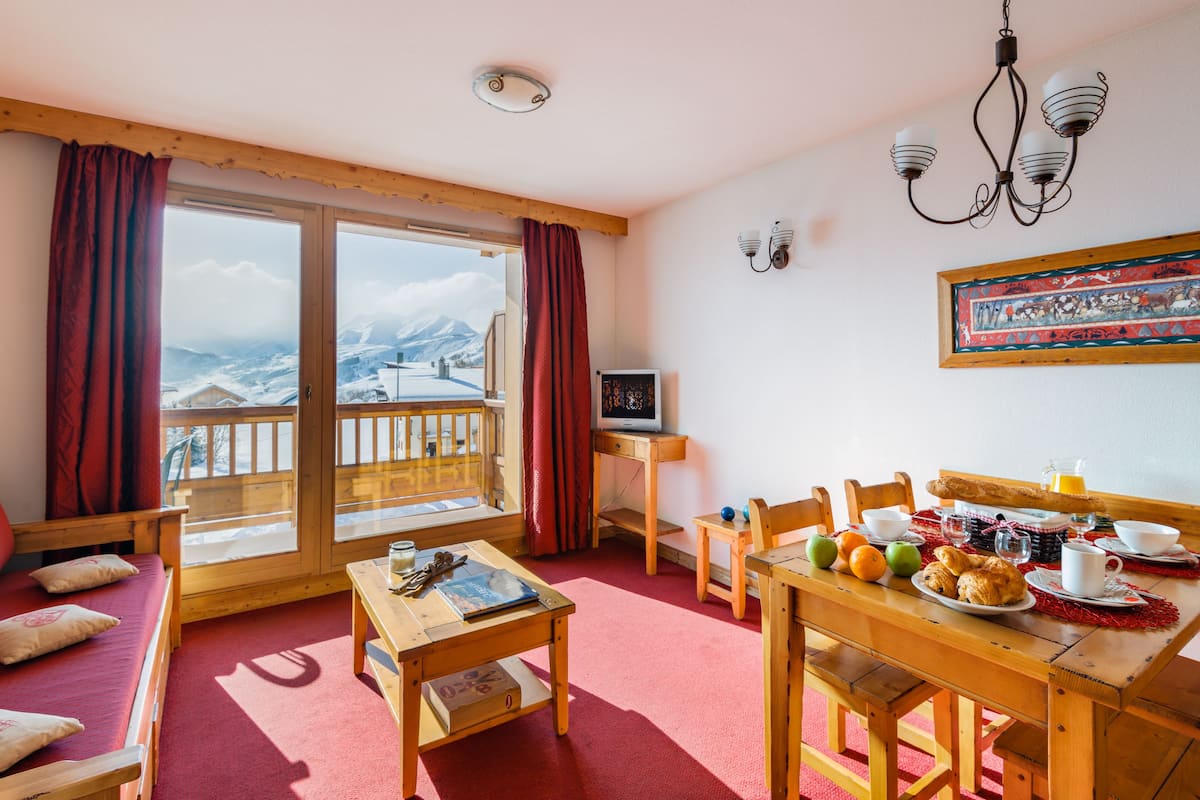 Ein gemütliches Zimmer mit einem hölzernen Bett, einem kleinen Tisch mit Frühstücksangeboten, einem Fernseher und einem Balkon mit Bergblick.