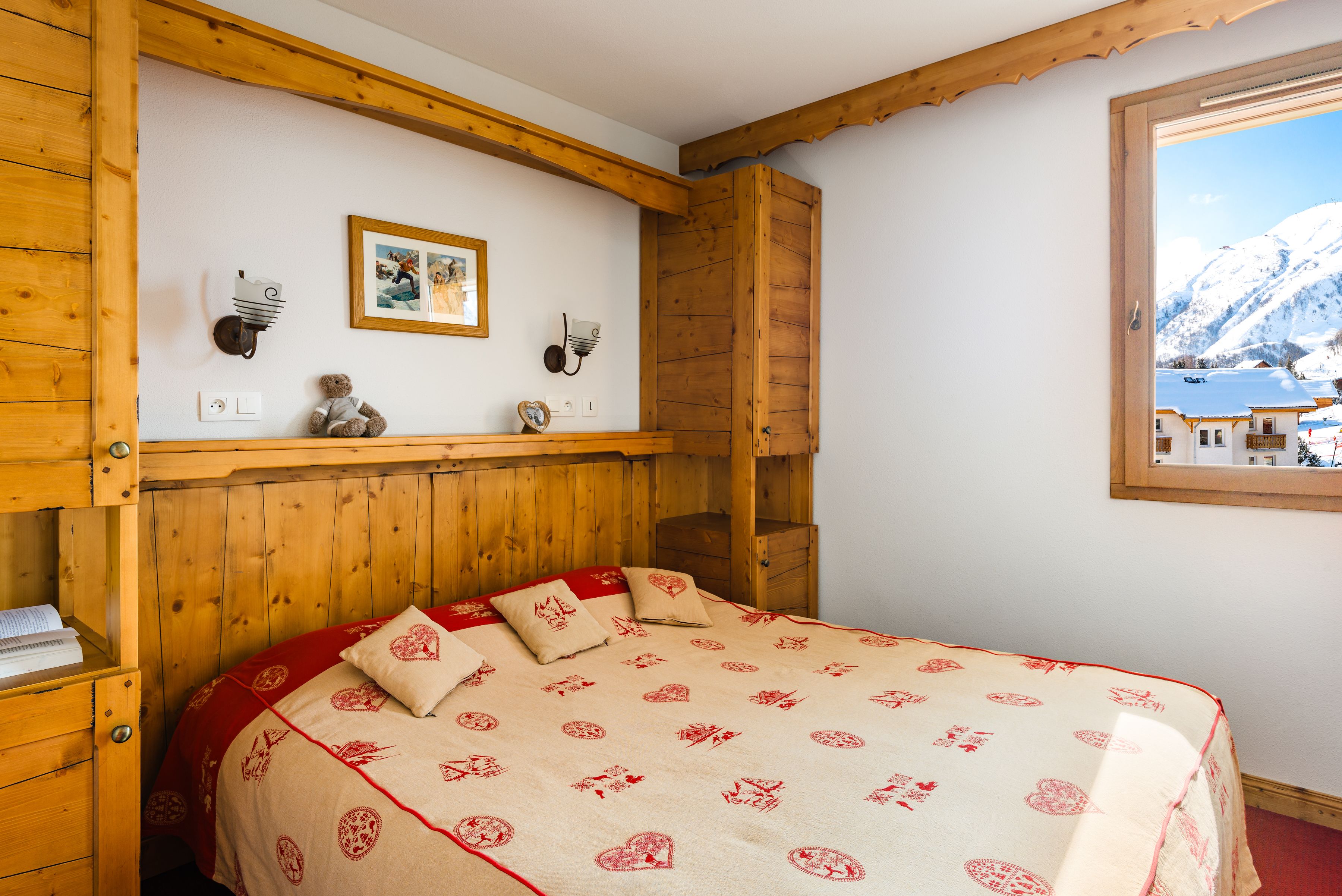 Ein gemütliches Zimmer mit einem hölzernen Bett, einer kleinen Ablage mit Dekoration und einem Fenster mit Blick auf einen verschneiten Berg.