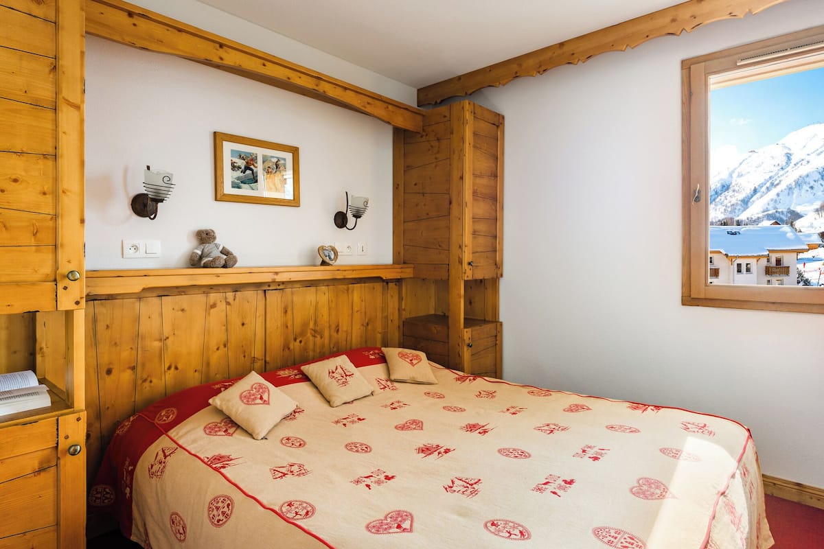 Ein gemütliches Zimmer mit einem Holzkopfteil, einem Bett mit einer rot-weißen Bettdecke, einer kleinen Ablagefläche mit Dekoration und einem Fenster mit Blick auf einen verschneiten Berg.