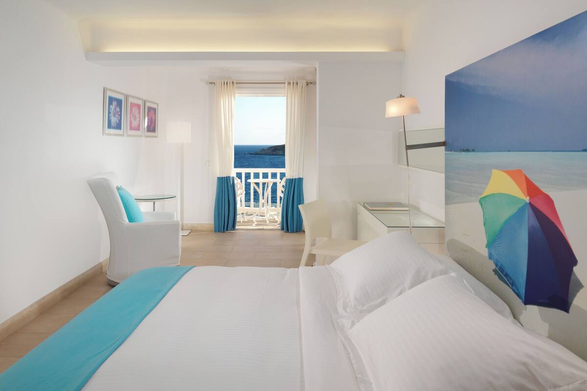 A hotel room with a bed, a desk, a chair, a balcony with curtains, and a painting of a beach.