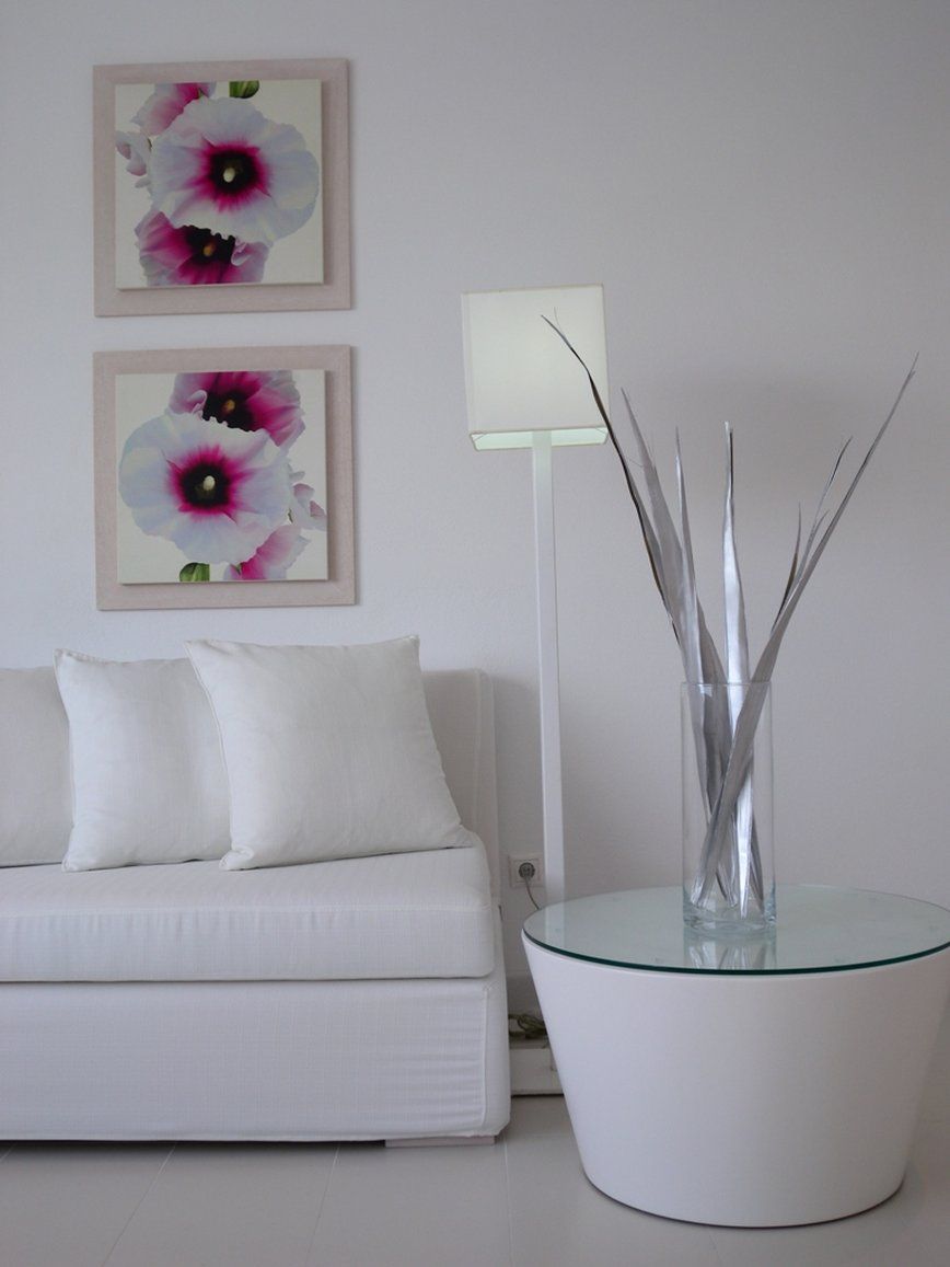 A white sofa with two cushions, a glass table with a vase, and two framed floral paintings on the wall.