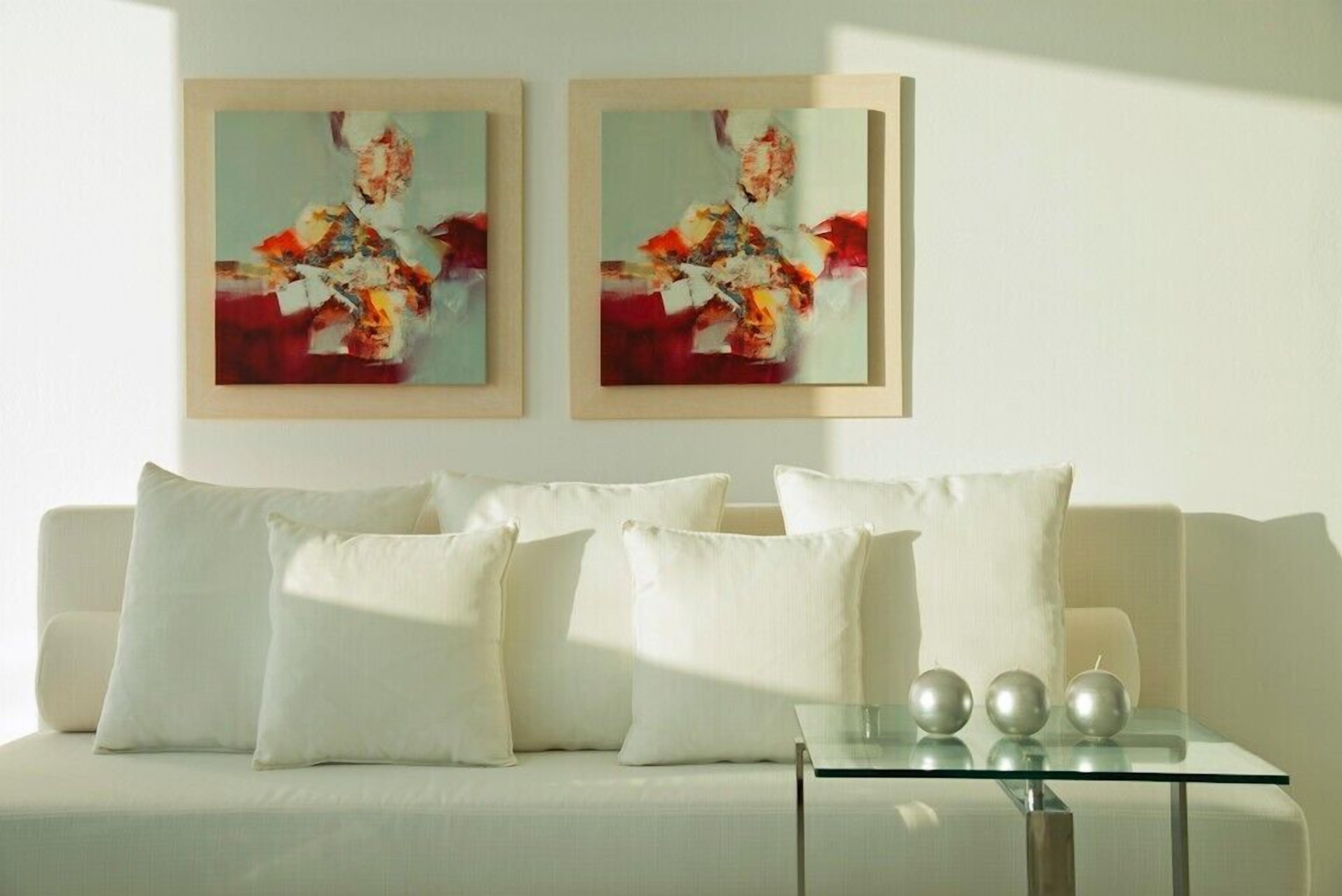 Ein modernes Wohnzimmer mit einem weißen Sofa, drei Kissen und einem Glastisch mit drei metallischen Kugeln. Über dem Sofa hängen zwei gerahmte abstrakte Gemälde.