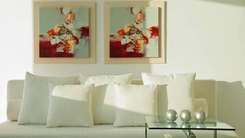 Ein modernes Wohnzimmer mit einem weißen Sofa, drei Kissen und einem Glastisch mit drei metallischen Kugeln. Über dem Sofa hängen zwei gerahmte abstrakte Gemälde.