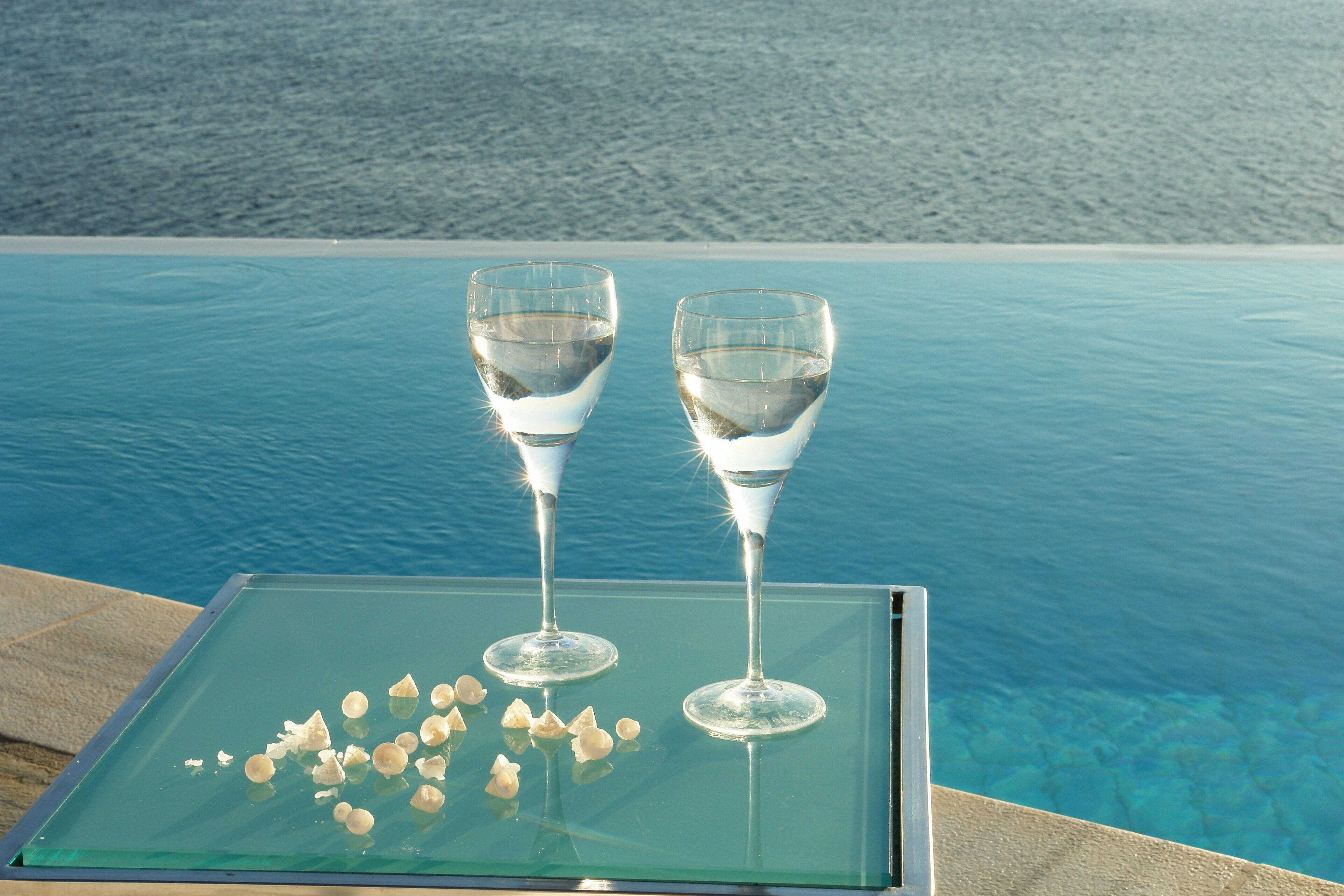 Two glasses of water on a glass table by a pool with a beach view.
