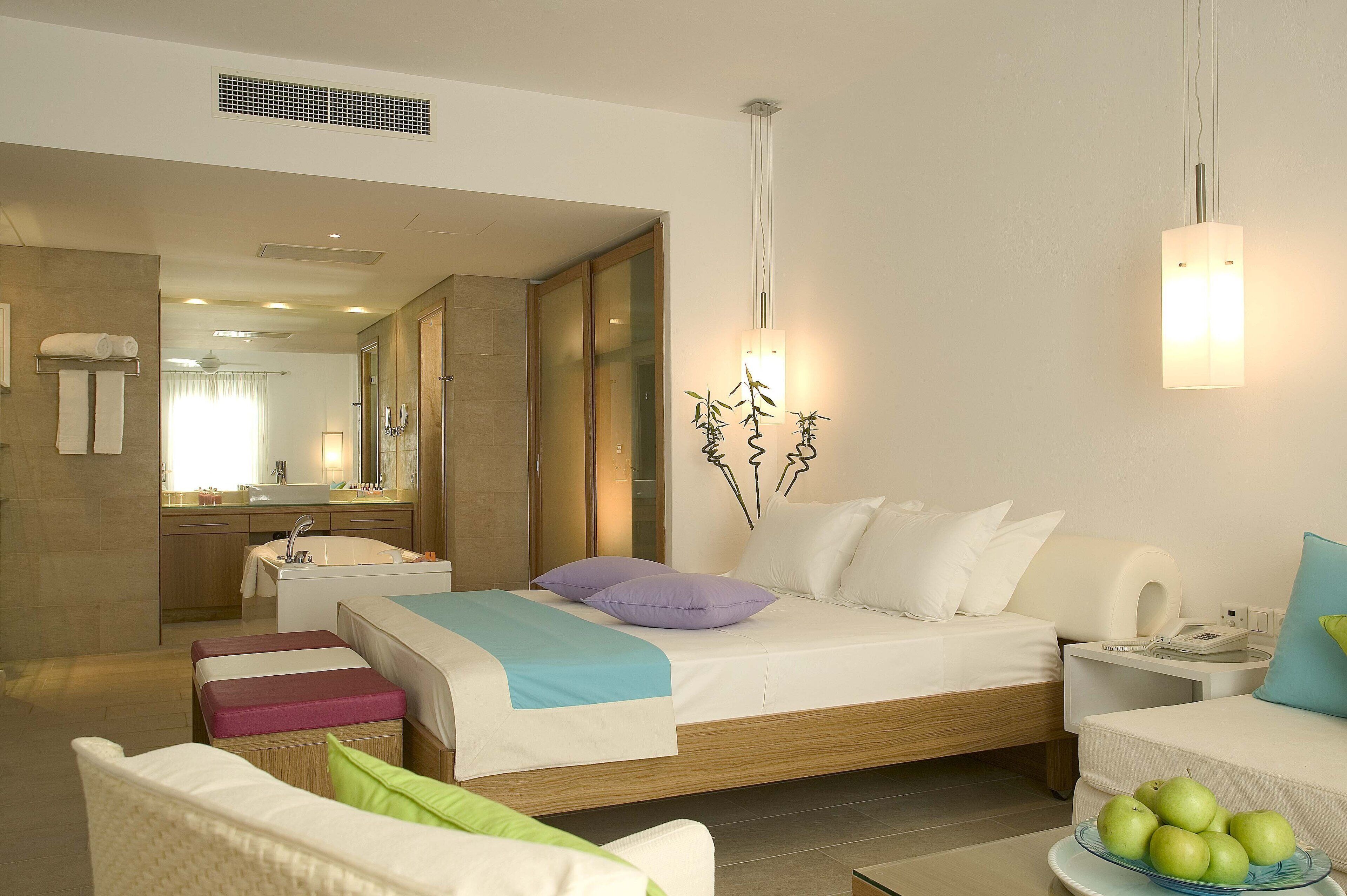A modern hotel room with a large bed, a sitting area with a chair and ottoman, a small table with fruit, and a bathroom visible through an open door.
