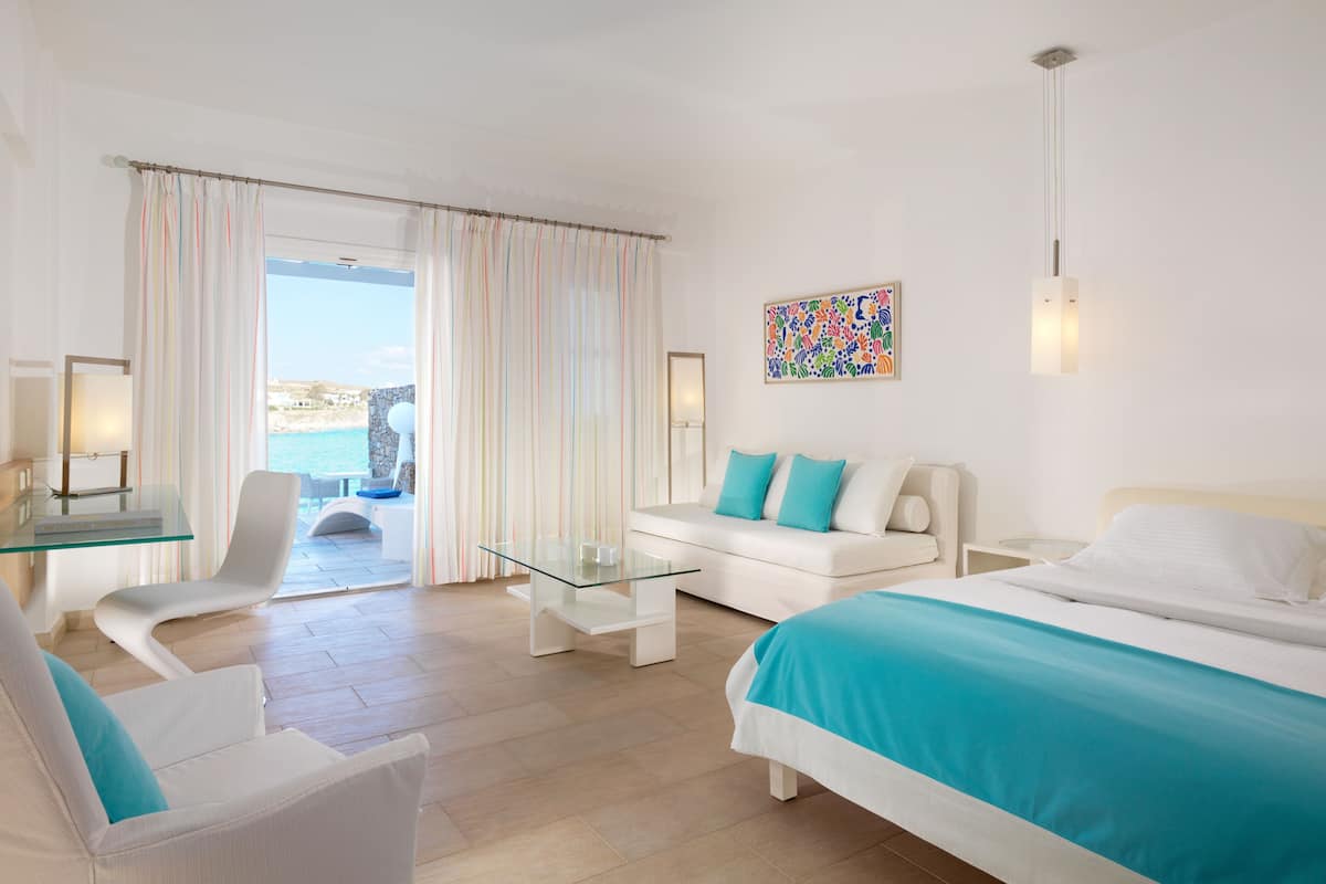 A modern hotel room with a bed, a sofa, a glass coffee table, a chair, and a view of the sea.