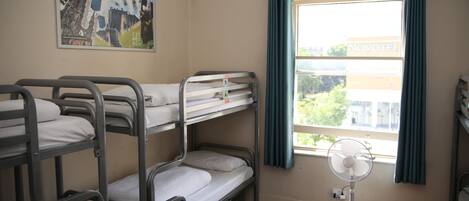 A dormitory room with bunk beds, a window with curtains, a fan, and a poster on the wall.
