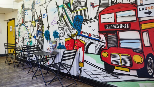 A room with a mural of London landmarks, a red double-decker bus labeled 'Greenwich', and foldable chairs. - St Christopher's Inn, Greenwich - Hostel (London)